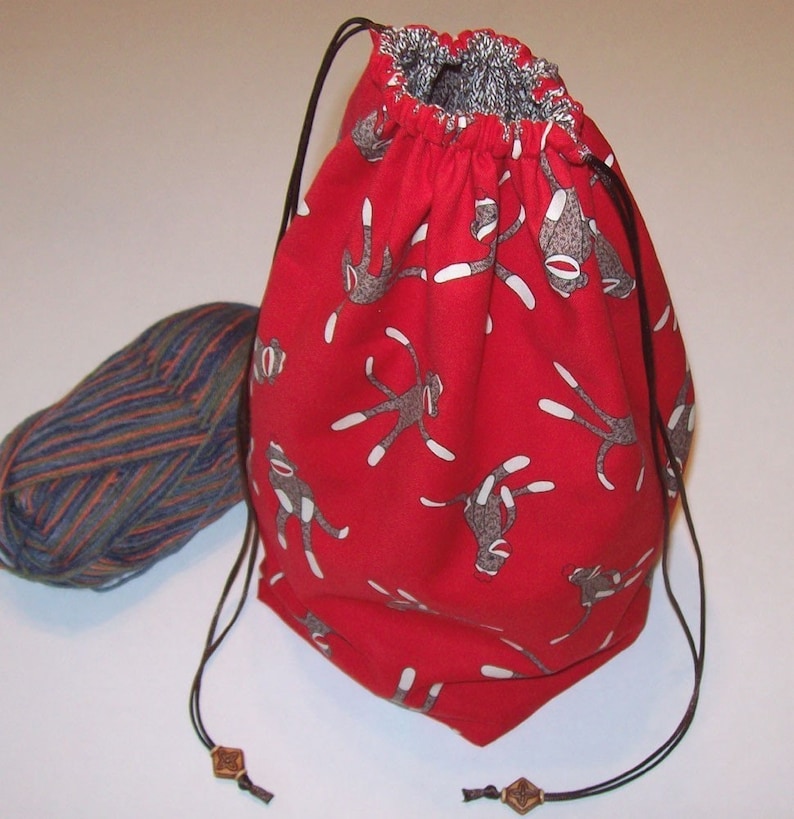K&ouml;nnte beinhalten: Rote Kordeltasche mit Affen-Motiv. Die Tasche ist aus Stoff und hat einen Kordelverschluss. Die Tasche ist leer.