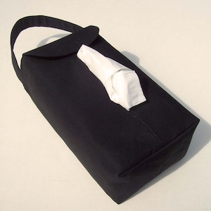 May include: A black fabric tissue box holder with a handle. The holder has a flap that opens to allow access to the tissues. A white tissue is partially visible through the opening.
