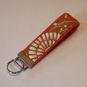 May include: A fabric keychain wristlet with a floral pattern in shades of orange, white, and green. The wristlet is bordered with a solid orange fabric and includes a silver metal key ring.