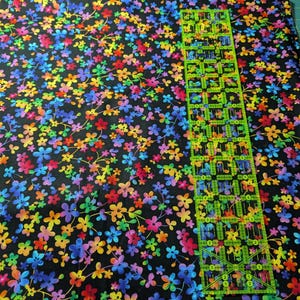 May include: A piece of fabric with a black background covered in a vibrant floral pattern. The flowers are in shades of red, orange, yellow, green, blue, and purple. A clear, green ruler is placed on top of the fabric, showing inch measurements.