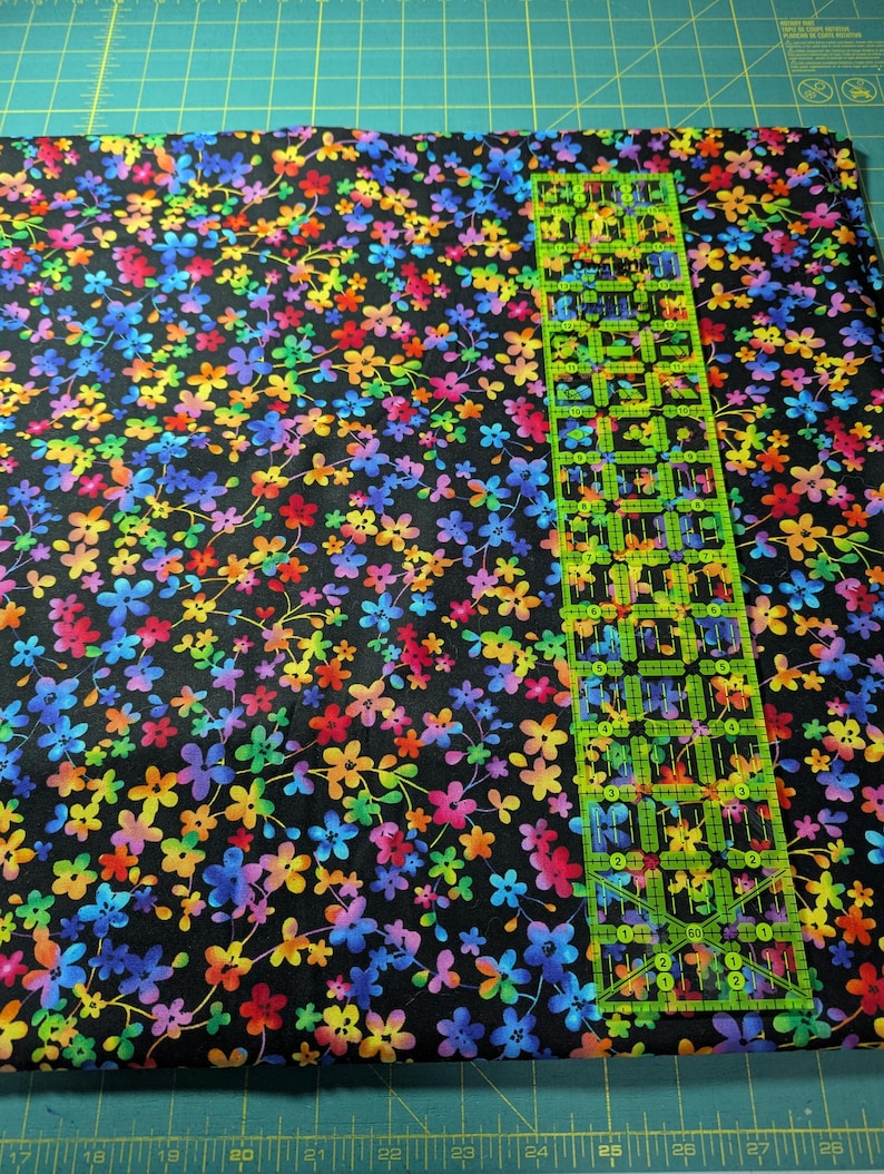 May include: A piece of fabric with a black background covered in a vibrant floral pattern. The flowers are in shades of red, orange, yellow, green, blue, and purple. A clear, green ruler is placed on top of the fabric, showing inch measurements.