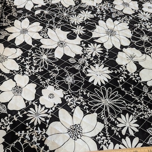 May include: Black and white floral quilted fabric. Large white flowers and smaller floral designs are set against a black background. The fabric is quilted with a diamond pattern. A wooden ruler is at the bottom of the image.