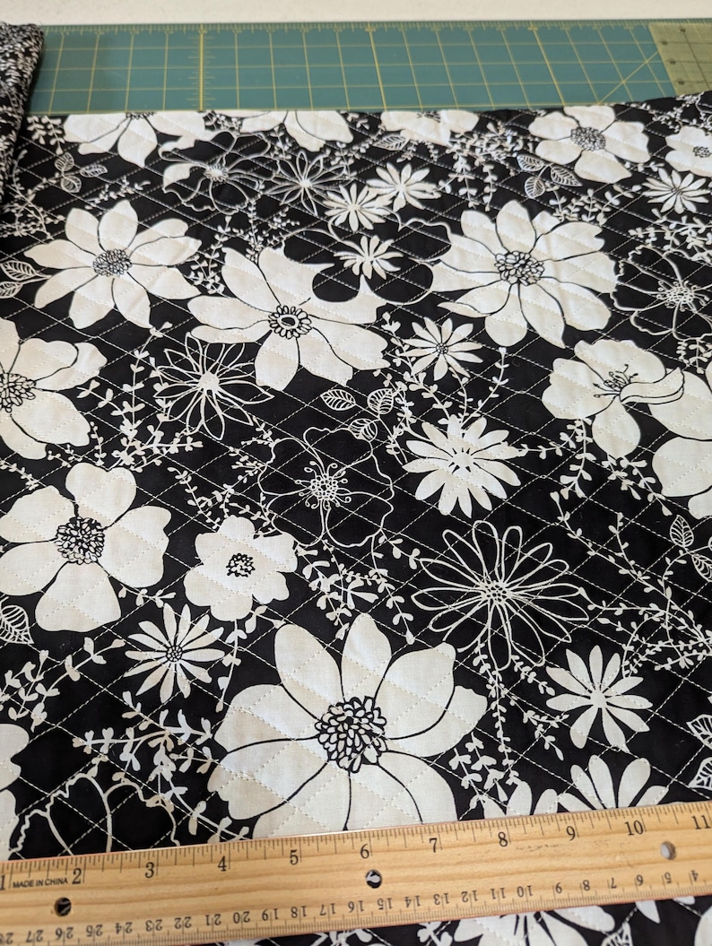 May include: Black and white floral quilted fabric. Large white flowers and smaller floral designs are set against a black background. The fabric is quilted with a diamond pattern. A wooden ruler is at the bottom of the image.