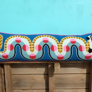 May include: A long, blue pillow with a pink, yellow, and gray geometric pattern. The pattern features a series of circles and wavy lines.
