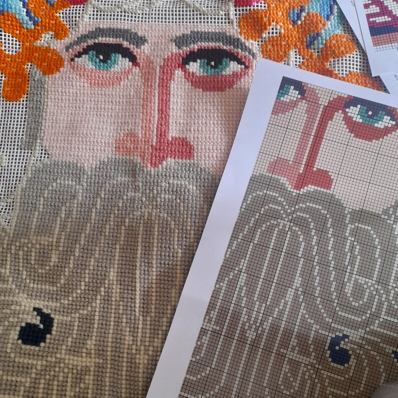 K&ouml;nnte beinhalten: Nahaufnahme eines Kreuzstichprojekts mit einem stilisierten Gesicht mit rosa Nase und Lippen, blauen Augen und einem grauen Bart. Das Design umfasst orangefarbene und gelbe florale Akzente. Ein gedrucktes Muster liegt daneben.