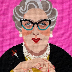 May include: A needlepoint portrait of a woman with gray hair, wearing turquoise glasses, a pearl necklace, and a black dress. She is holding a pair of scissors in her hand.