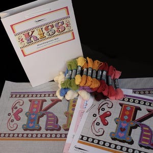 May include: A needlepoint kit with colorful embroidery floss, a canvas with a pre-printed design of the word "KISS" in large letters, and instructions. The kit is packaged in a white bag with the word "KISS" printed on it.