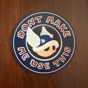 May include: A round sticker with a blue and white design. The sticker features a cartoon character wearing a blue helmet with white wings. The text "DON'T MAKE ME USE THIS" is printed around the character in red and white.
