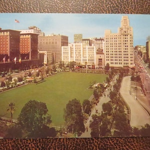 May include: A vintage postcard depicting a city park with tall buildings in the background. The park is green and has trees and a path. The buildings are a mix of brick and concrete and have a variety of architectural styles.