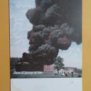 May include: A vintage postcard depicting a large plume of black smoke rising from a burning oil tank in Olean, NY. The smoke billows high into the sky, obscuring the surrounding area. Several people are standing in the foreground, watching the fire.