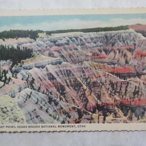 May include: Vintage postcard featuring Sunset Point in Cedar Breaks National Monument, Utah. The image showcases a colorful canyon landscape with layers of red, white, and purple rock formations. The top of the postcard has a line of green trees and a blue sky.