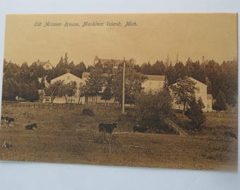May include: Sepia-toned postcard featuring the Old Mission House on Mackinac Island, Michigan. The image shows a large white building surrounded by trees, with cows grazing in a field in the foreground. The text "Old Mission House, Mackinac Island, Mich." is printed at the top.