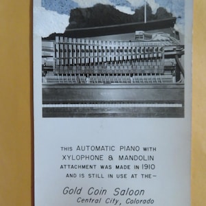 May include: Black and white photo of an automatic piano with a xylophone and mandolin attachment. The text on the postcard reads: "This automatic piano with xylophone & mandolin attachment was made in 1910 and is still in use at the - Gold Coin Saloon Central City, Colorado."