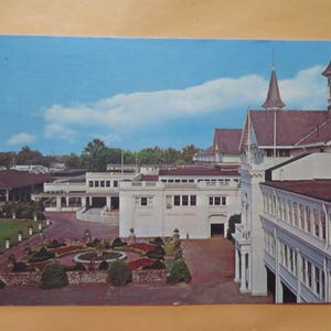 May include: A vintage postcard depicting a large white building with multiple towers and a red roof. The building is surrounded by a landscaped courtyard with trees and shrubs. The postcard is titled "Churchill Downs" in the upper right corner.