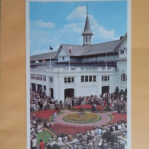 May include: A vintage postcard featuring a white building with a tall spire, surrounded by a circular flower bed and a crowd of people. The text on the postcard reads "Clubhouse Grounds at Churchill Downs".