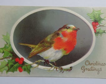 May include: A vintage Christmas postcard showing a robin with a red breast, perched on a branch inside an oval frame. The card features holly with red berries and the text "Christmas Greetings".