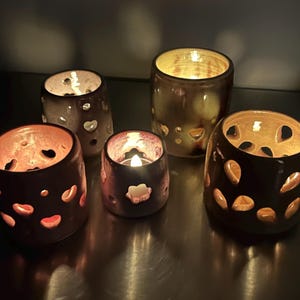 May include: Five ceramic candle holders in varying sizes and shapes. The holders are dark brown with heart-shaped cutouts, and some have a golden interior. A lit candle is inside one of the holders, casting a warm glow.