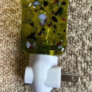 May include: A decorative night light with a rectangular, yellow-green glass face. The glass is embedded with colorful specks of white, black, and red. The base is white with a two-prong plug.