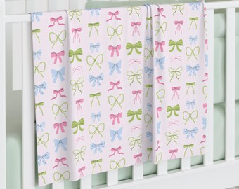 May include: A baby blanket with a repeating pattern of bows in pink, blue, and green on a light pink background. The blanket is draped over a white crib.