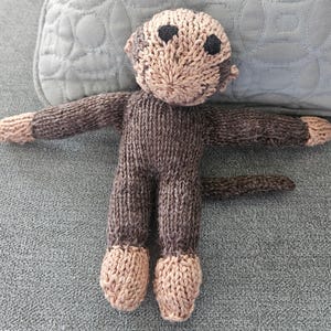 May include: A handmade knitted monkey doll in shades of brown and tan. The monkey has a detailed face with black eyes and a textured body. The arms and legs are extended, and the tail is curled. The doll is made of soft yarn, perfect for a child's toy or nursery decor.