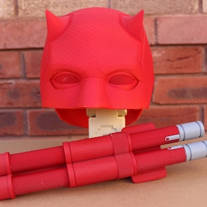 May include: A red superhero mask with horns and eye mesh, paired with two red and gray batons. The mask is the focal point, with the batons resting below. The items are set against a brick wall.