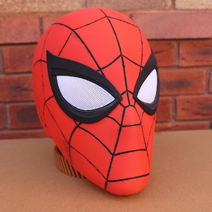 May include: A red Spider-Man mask with black webbing and large white eye lenses outlined in black. The mask is made of a textured fabric and sits on a brown base. The background features a red brick wall.