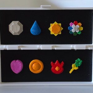 Könnte beinhalten: Ein weißes rechteckiges Etui mit einer schwarzen Schaumstoffeinlage, das acht kleine, bunte, dreidimensionale Formen enthält. Die Sammlung umfasst ein graues Achteck, einen blauen Tropfen, eine gelbe Sonne, eine mehrfarbige Blume, ein rosa Herz, einen orangefarbenen Kreis, eine rote Flamme und ein grünes pflanzenartiges Symbol.