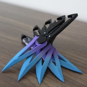 Könnte beinhalten: Ein Satz von sechs Wurfmessern mit schwarzem Griff und blauen und violetten Ombre-Klingen. Die Messer sind in einem Sternmuster angeordnet.