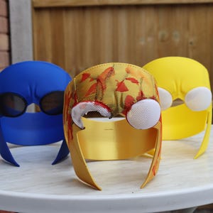 Peut inclure: Trois masques de costume colorés. Un masque bleu avec des couvre-yeux en maille noire, un masque jaune et un masque jaune avec un motif floral. Les masques sont sur une table blanche.