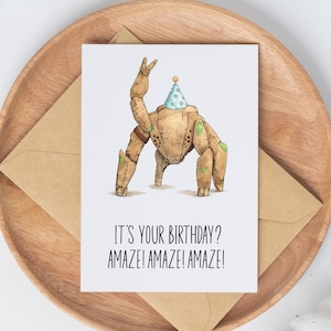 May include: A white greeting card featuring a brown, stone-like robot character wearing a light blue polka-dot party hat. The robot is making a peace sign with one hand. Below the illustration, the text reads: IT'S YOUR BIRTHDAY? AMAZE! AMAZE! AMAZE!