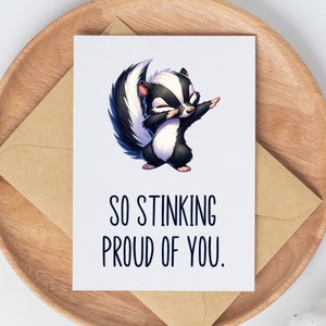 May include: A white card with a cartoon skunk in a dabbing pose. The text on the card reads "So Stinking Proud of You."