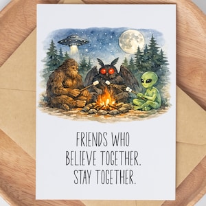 May include: A greeting card with an illustration of Bigfoot, a Mothman, and an alien roasting marshmallows around a campfire under a full moon. The card reads "FRIENDS WHO BELIEVE TOGETHER. STAY TOGETHER." The card is on a wooden tray with a brown envelope.