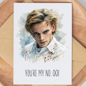 May include: Greeting card with a watercolor portrait of a person with blonde hair and a stern expression. The card reads "YOU'RE MY NO. 001" in black text. The card is on a wooden plate with a brown envelope.
