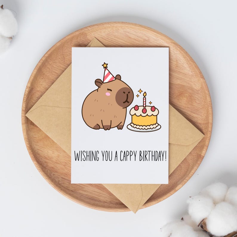 May include: A birthday greeting card featuring a cartoon capybara wearing a party hat next to a cake with a lit candle. The card is on a wooden tray with a brown envelope. The text on the card reads, "WISHING YOU A CAPPY BIRTHDAY!"