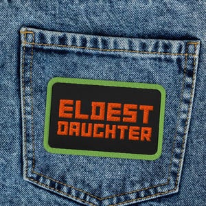 Könnte beinhalten: Nahaufnahme einer hellblauen Jeans mit einer Gesäßtasche. Ein rechteckiger Aufnäher mit den Worten "ELDEST DAUGHTER" in orangefarbenen Buchstaben ist auf die Tasche genäht. Der Aufnäher hat einen schwarzen Hintergrund und einen grünen Rand.