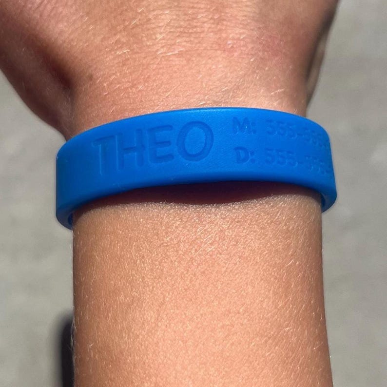 May include: A blue silicone wristband with the name "THEO" embossed on the front. The wristband also displays a phone number and a date. The wristband is worn on a wrist.