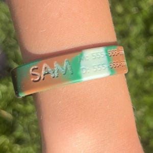 May include: A camouflage-patterned silicone wristband with the name "SAM" in white letters. The wristband also displays two phone numbers. The color scheme includes shades of green, brown, and white.