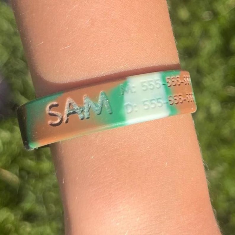 May include: A camouflage-patterned silicone wristband with the name "SAM" in white letters. The wristband also displays two phone numbers. The color scheme includes shades of green, brown, and white.