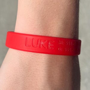 May include: A red silicone wristband with the name "LUKE" embossed on the front. Additional text and numbers are visible on the band. The wristband is worn on a person's wrist.