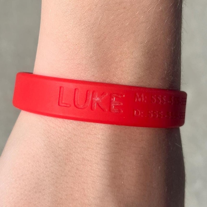 May include: A red silicone wristband with the name "LUKE" embossed on the front. Additional text and numbers are visible on the band. The wristband is worn on a person's wrist.