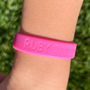 May include: A bright pink silicone wristband with the name "RUBY" embossed on the front. The wristband also has two lines of text with phone numbers. The background is blurred green.