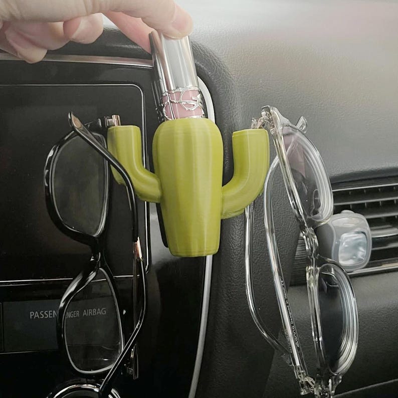 May include: A green cactus-shaped car organizer with two arms holding glasses and a lipstick tube in the center.