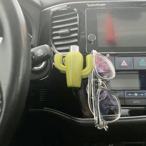 May include: A green cactus-shaped car air freshener hanging from a car's rearview mirror. A pair of glasses is also hanging from the mirror.