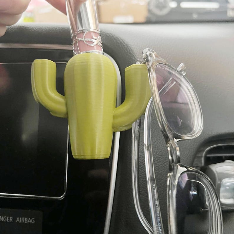 May include: A green cactus-shaped car accessory with a lipstick and glasses holder. The cactus is made of plastic and has a textured surface.