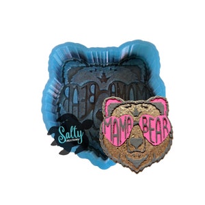 May include: A silicone mold for making resin crafts. The mold is shaped like a bear wearing heart-shaped sunglasses and the text "Mama Bear".
