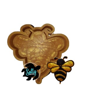 May include: A golden bee-shaped mold and a bee-shaped charm. The mold is large and detailed, with a textured surface. The charm is black and gold with striped detailing. The logo for Salty Creations is in the lower left corner.
