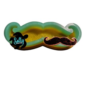 May include: A green, yellow, and orange resin mustache mold with a black mustache design. The mold is made by Salty Creations.
