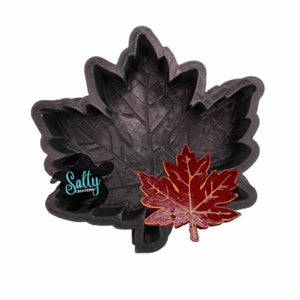 May include: A black silicone mold shaped like a maple leaf. A red maple leaf is sitting next to the mold. The mold is for making resin crafts.