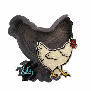 May include: A white chicken with a red comb and wattle, standing on a black silicone mold of a chicken. The mold is textured and has a raised design of a chicken.