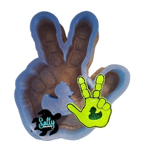 May include: A peace sign-shaped mold with a duck-shaped cutout. The mold is brown with blue edges. A neon green peace sign with a duck graphic is next to the mold. The text "Salty Creations" is on the bottom left.